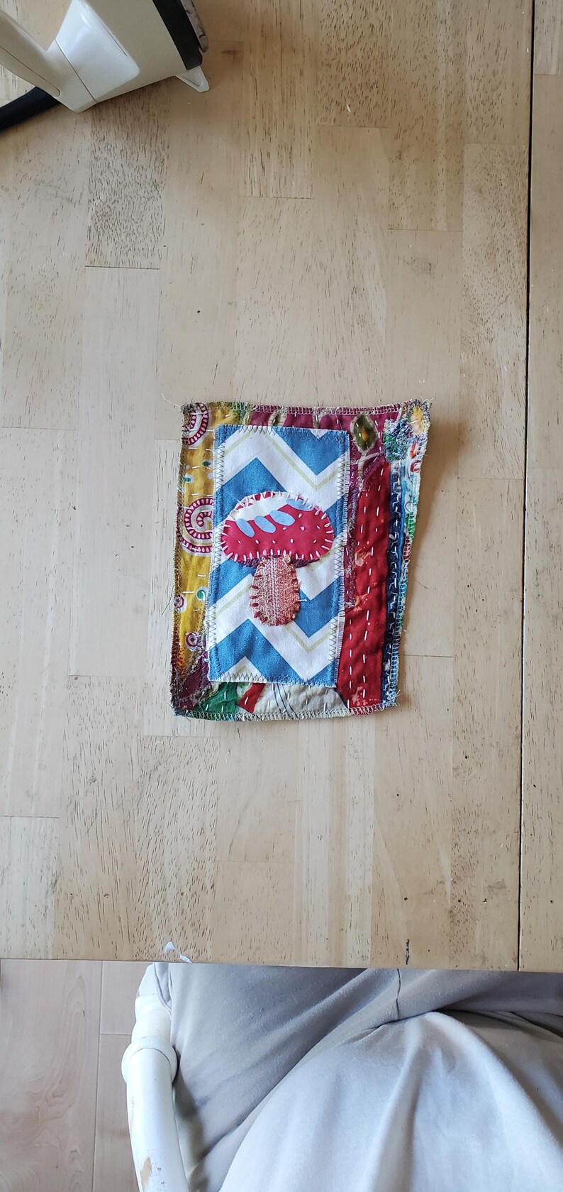 May include: A handmade fabric patch with a mushroom applique. The patch features a red mushroom with a white and blue chevron pattern, and is bordered by various patterned fabrics.