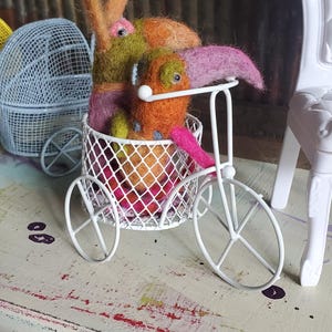 May include: A charming scene with felted bunny figures in a white wire bicycle basket. The bunnies are in shades of orange, green, and pink. A white wire stroller and chair are in the background, set on a distressed, multi-coloured surface.