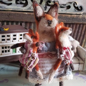 May include: A felted fox family figurine. The mother fox, with orange and white fur, wears a blue and white apron and holds two smaller foxes. The small foxes wear dresses in pink and red patterns. The background includes a miniature house and furniture.