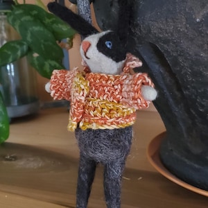 May include: A black and white felt rabbit wearing a crocheted orange and yellow sweater. The rabbit is standing on a wooden surface.