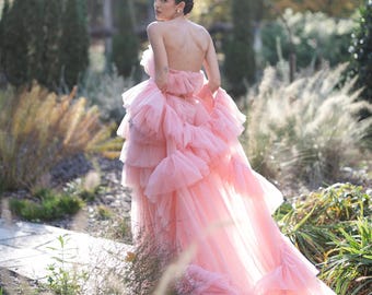 May include: A woman wearing a pink tulle gown with multiple layers of ruffles. The gown has a long train and is strapless. The woman is standing in a garden setting.