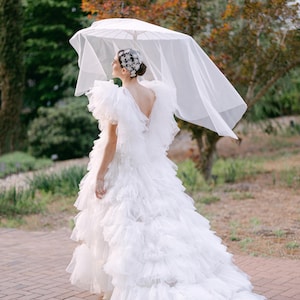 May include: A white wedding dress with a layered, ruffled skirt and a low back. The dress has a long train and is paired with a sheer veil and a decorative headpiece. The person is holding a white parasol.