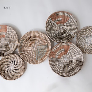 Puede incluir: Un conjunto de seis cestas de pared redondas tejidas etiquetadas como Set B. Las cestas presentan dise&ntilde;os en espiral en tonos neutros de beige, blanco, terracota y marr&oacute;n oscuro, con varios patrones geom&eacute;tricos tejidos en fibra natural.