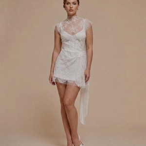 May include: A white lace mini dress with a high neckline and cap sleeves. The dress features a fitted bodice and a flowing skirt with a side drape. The model is wearing white heels.