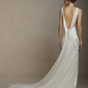 May include: A white wedding dress with a low back and a long, flowing train. The dress has a simple, elegant design with thin straps and a draped skirt. The back of the dress is open, revealing the wearer's back. The train extends across the floor.