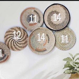 Puede incluir: Una colecci&oacute;n de seis cestas de pared tejidas redondas con fibras naturales en tonos bronceado, marr&oacute;n, negro y azul verdoso apagado. Las cestas est&aacute;n dispuestas en grupo, cada una marcada con un n&uacute;mero blanco 11, 12 o 14, con espirales.