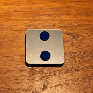 May include: A square, brushed metal object with two evenly spaced, circular, dark blue insets. The object is set against a warm, wooden background.
