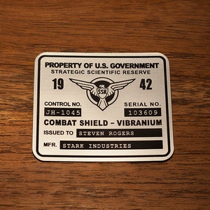 May include: A silver metal tag with black text that reads "PROPERTY OF U.S. GOVERNMENT STRATEGIC SCIENTIFIC RESERVE 19 42 CONTROL NO. JH-1045 SERIAL NO. 103609 COMBAT SHIELD - VIBRANIUM ISSUED TO STEVEN ROGERS MFR. STARK INDUSTRIES". The tag features a black and white image of a winged shield with the letters "SSR" in the center.