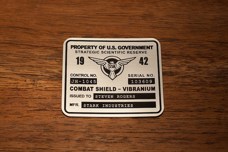 May include: A silver metal tag with black text that reads "PROPERTY OF U.S. GOVERNMENT STRATEGIC SCIENTIFIC RESERVE 19 42 CONTROL NO. JH-1045 SERIAL NO. 103609 COMBAT SHIELD - VIBRANIUM ISSUED TO STEVEN ROGERS MFR. STARK INDUSTRIES". The tag features a black and white image of a winged shield with the letters "SSR" in the center.