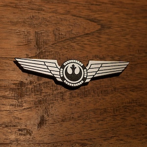 May include: A silver Rebel Alliance pilot wing pin with a black and white symbol in the centre. The pin is on a brown wooden surface.