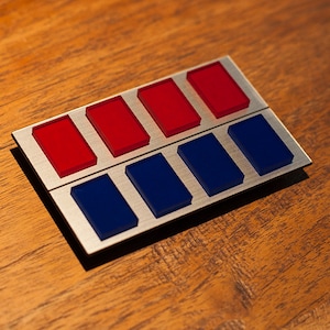 May include: A silver metal badge with eight rectangular red and blue colored insets. The badge is on a wooden surface.
