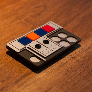 Könnte beinhalten: Eine kleine, rechteckige graue Box mit einer silbernen Frontplatte. Die Frontplatte hat vier farbige Knöpfe: rot, orange, blau und blau. Außerdem befinden sich sechs runde, silberfarbene Knöpfe an der Box.
