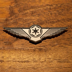 May include: A white and black metal pin with a winged design and a star-shaped symbol in the center. The pin is on a wooden surface.