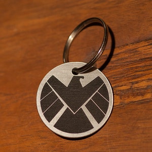 May include: A silver key chain with a round metal tag featuring a black and white logo of a shield with a bird inside. The shield has a textured surface.