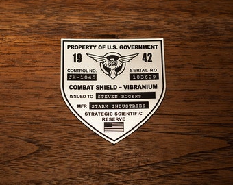 May include: A silver shield with black text and a black eagle logo. The text reads "PROPERTY OF U.S. GOVERNMENT", "CONTROL NO. JH-1045", "SERIAL NO. 103609", "COMBAT SHIELD - VIBRANIUM", "ISSUED TO STEVEN ROGERS", "MFR STARK INDUSTRIES", "STRATEGIC SCIENTIFIC RESERVE".