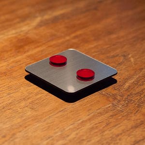 May include: A square, brushed metal object with two small, round, red pieces on top. The object is set on a wooden surface. The red pieces are evenly spaced on the metal square.
