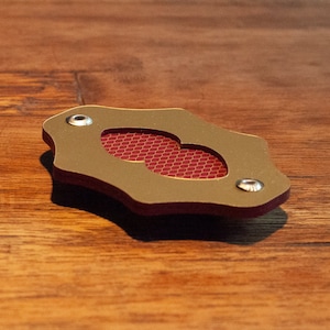 May include: A gold-coloured metal object with a red honeycomb pattern inside. The object has a unique shape and three silver screws.