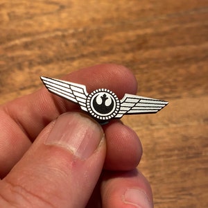 Op de afbeelding: Een zilveren pin met een zwart-wit Rebel Alliance-symbool en vleugels. De pin is een populair verzamelstuk voor Star Wars-fans.