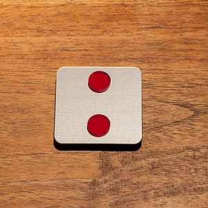 May include: A square, silver-colored object with two red circular accents, one above the other. The object is set against a wooden background. The design is simple and modern.