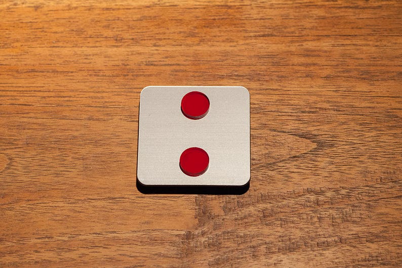 May include: A square, silver-colored object with two red circular accents, one above the other. The object is set against a wooden background. The design is simple and modern.