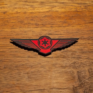 Könnte beinhalten: Eine schwarze und rote Anstecknadel mit einem geflügelten Design und dem imperialen Symbol aus Star Wars.