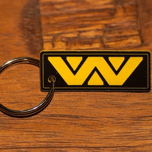 May include: A rectangular keychain featuring a black background with a bold, yellow geometric design resembling the letters W and V. The keychain includes a circular silver metal split ring attached to the left side of the rectangular tag.