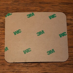 May include: A brown square adhesive sheet with the 3M logo printed in green on it.