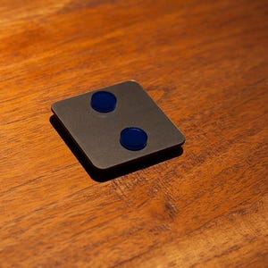 May include: A square, dark gray object with two circular, blue accents. The object is set on a wooden surface with a warm, brown tone. The lighting creates a shadow on the surface.