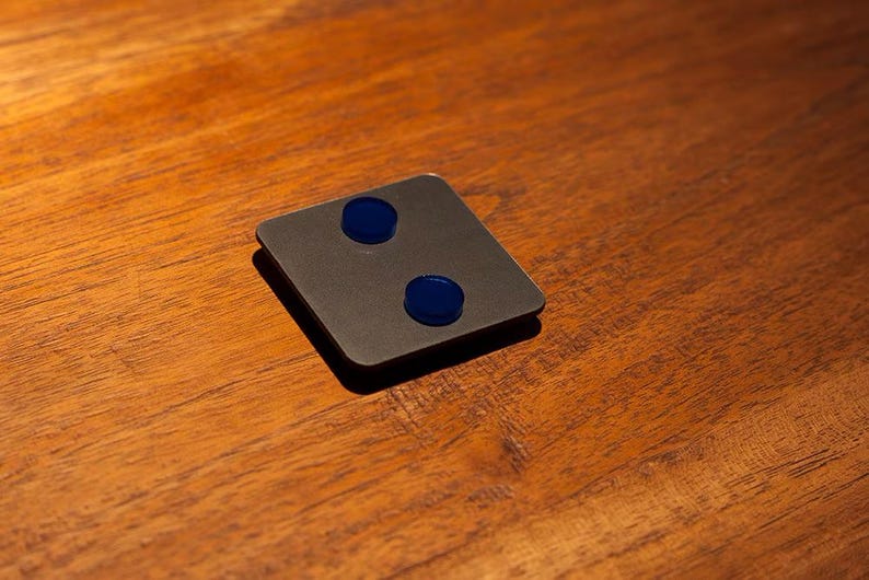 May include: A square, dark gray object with two circular, blue accents. The object is set on a wooden surface with a warm, brown tone. The lighting creates a shadow on the surface.