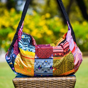 May include: A vibrant patchwork shoulder bag with a black strap. The bag displays a variety of patterned fabrics in shades of blue, yellow, red, and purple. The bag is resting on a woven basket.