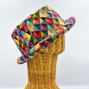 Può includere: Un cappello fedora colorato con un motivo geometrico di triangoli in vari colori, tra cui rosso, giallo, blu e verde. Il cappello è esposto su una testa di manichino in vimini, che ne mette in risalto la forma e il design.