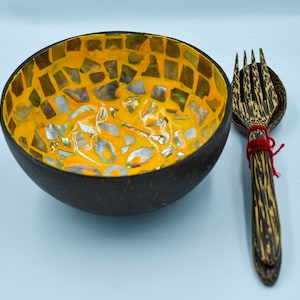 May include: A handcrafted coconut bowl with a mosaic interior of yellow, silver, and iridescent tiles. A wooden spoon and fork set, tied with red twine, rests beside the bowl. The bowl is dark brown on the outside.