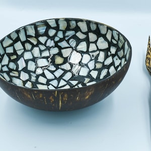 May include: A decorative coconut bowl with a mosaic shell interior and a set of wooden utensils. The bowl is brown with a mosaic pattern of white and gray shells. The utensils are tied together with red twine.