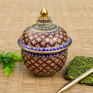May include: A round ceramic lidded jar featuring a deep red base colour with intricate hand-painted floral patterns in white, blue, and gold. The lid is topped with a pointed gold finial, and the entire surface is decorated with detailed geometric motifs and vibrant enamel colours, showcasing a traditional craftsmanship style with a glossy finish.