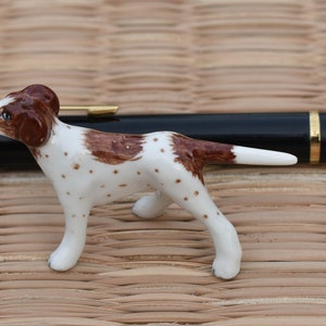 Può includere: Una piccola statuetta di porcellana di un cane macchiato di marrone e bianco. Il cane è in piedi su un tappetino intrecciato e guarda a sinistra.