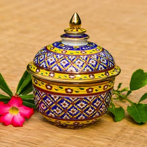 Op de afbeelding: Een decoratieve keramische kom met deksel, met een levendig blauw, geel en wit bloemmotief. De kom heeft een goudkleurige rand en een gouden knop op het deksel. Geplaatst op een geweven oppervlak met een roze bloem en groene bladeren.