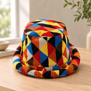 Puede incluir: Un sombrero de pescador con un patrón geométrico repetitivo y vibrante de triángulos en tonos brillantes de rojo, amarillo, azul, naranja y negro. El sombrero tiene una corona plana y un ala inclinada hacia abajo, hecho de tela tejida texturizada.