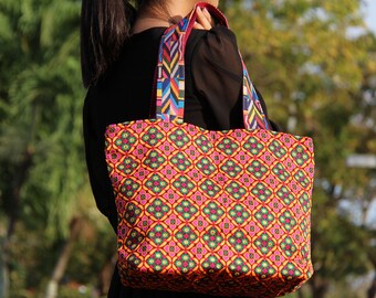 May include: A large tote bag with a colourful geometric pattern. The bag has a bright pink, yellow, green, and orange design. The straps are made of colourful woven fabric.