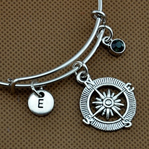 May include: A silver expandable bracelet with a compass charm, a letter 'E' charm, and a green gemstone charm.