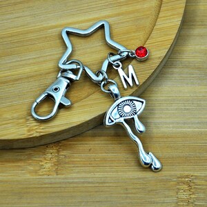 Puede incluir: Llavero de plata con un dije de estrella, un dije de la letra M, un dije de cristal rojo y un dije de plata de un ojo lloroso.