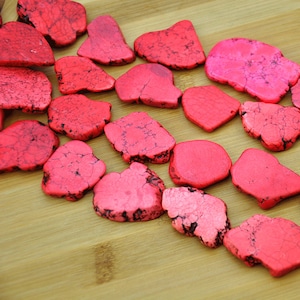 May include: A collection of pink and red dyed, natural, irregular shaped, smooth, polished, gemstone chips. The chips are arranged in a row on a wooden surface.