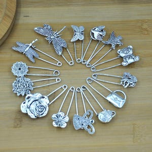 May include: A selection of silver-coloured safety pins, each adorned with a decorative charm. Charms include dragonflies, butterflies, flowers, a bird, a teddy bear, a purse, and a teapot. The pins are displayed on a wooden surface.