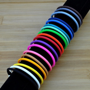 May include: A collection of thin, circular silicone bracelets in a spectrum of colors including black, white, yellow, orange, pink, purple, red, green, teal, blue, and brown, arranged in a row on a black cylindrical display stand.