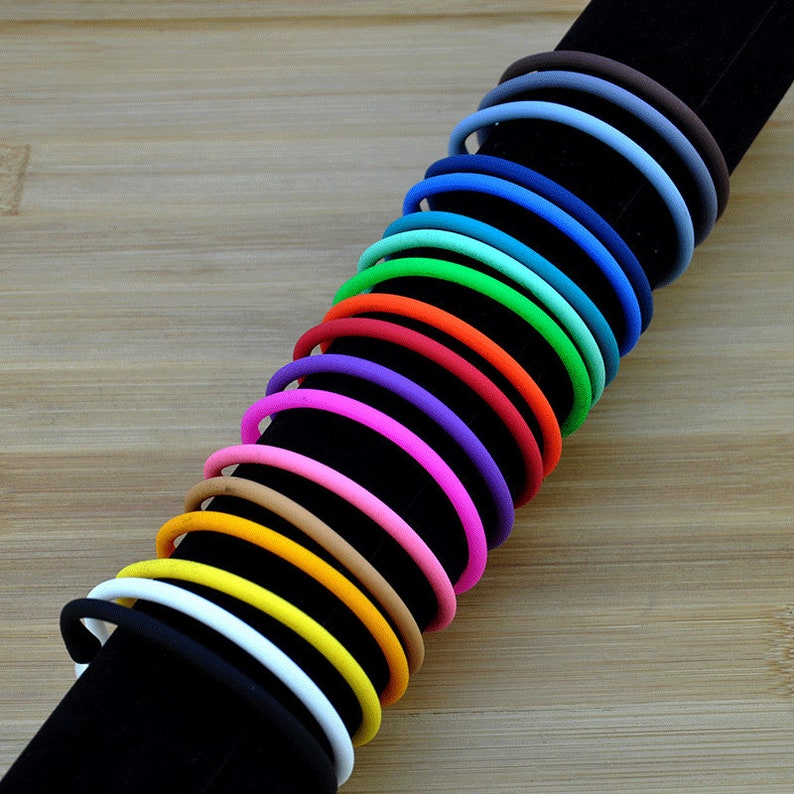 May include: A collection of thin, circular silicone bracelets in a spectrum of colors including black, white, yellow, orange, pink, purple, red, green, teal, blue, and brown, arranged in a row on a black cylindrical display stand.