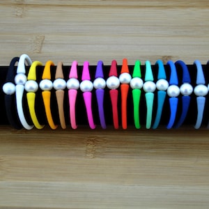 May include: A collection of fifteen flexible silicone bracelets arranged in a row, each featuring a single central white pearl bead. The bracelets come in a variety of solid colors including black, white, yellow, orange, tan, pink, purple, red, and blue.