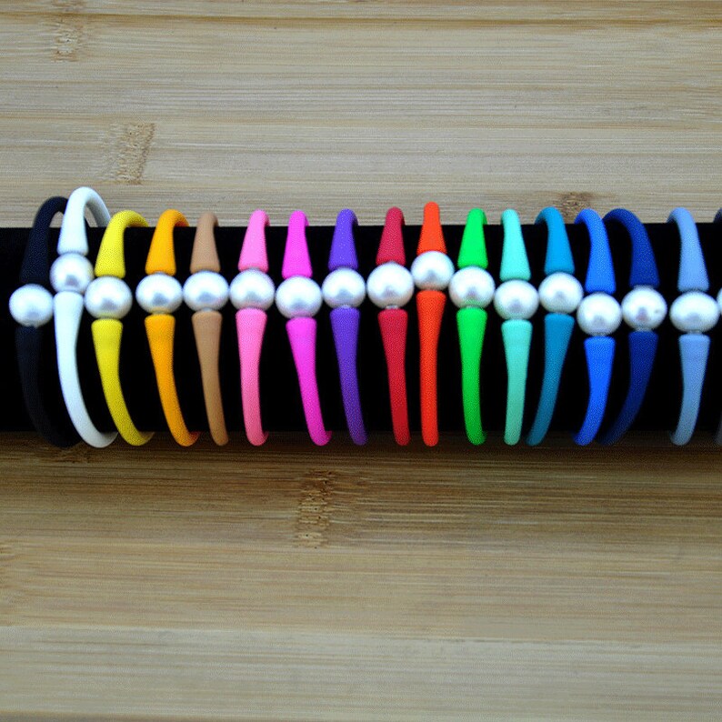 May include: A collection of fifteen flexible silicone bracelets arranged in a row, each featuring a single central white pearl bead. The bracelets come in a variety of solid colors including black, white, yellow, orange, tan, pink, purple, red, and blue.