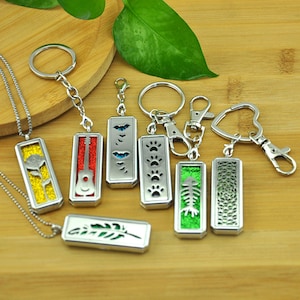 Puede incluir: Una colección de colgantes y llaveros rectangulares de metal plateado con diseños recortados que incluyen una rosa, guitarra, murciélagos, huellas, espina de pescado y patrones geométricos, con inserciones de purpurina de colores y cadenas plateadas.