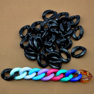 Pode incluir: Uma coleção de elos de corrente pretos em forma oval espalhados sobre uma superfície castanha, com uma corrente colorida de elos por baixo. A corrente colorida tem tons de azul, roxo, rosa e verde, com um elo de tartaruga na extremidade.