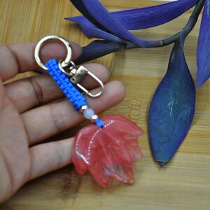 Puede incluir: Un llavero con un dije en forma de hoja roja. El dije está unido a un cordón azul tejido, un cierre plateado y un llavero plateado. El fondo incluye hojas moradas y una superficie de madera.