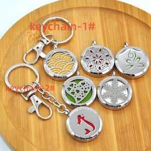 May include: Six silver keychains with different designs. Each keychain has a round metal locket with a different design, including a wave pattern, a snowflake, a heart, a flower, a leaf, and a fish. The keychains are on a wooden surface.